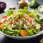 A vibrant Chinese Chicken Salad in a white bowl, featuring shredded chicken, crisp romaine lettuce, red bell peppers, shredded carrots, cucumber, and mandarin orange slices, topped with slivered almonds and sesame seeds. A light soy-ginger dressing drizzled over the salad, with chopsticks and a small bowl of dressing on a wooden table.